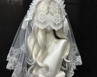 May include: A white lace headpiece featuring a ruffled headband with attached ribbons and a veil that drapes over the head. The veil includes intricate embroidered cross motifs and scalloped lace edges, resting on a white mannequin head.