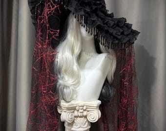 May include: A wide-brimmed black hat featuring ruffled fabric, hanging black beads, and a long black veil with a red spiderweb pattern. The hat is displayed on a white mannequin head with long white hair, which rests on a white classical column base.