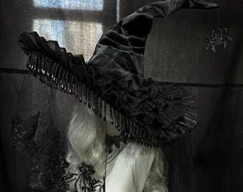 May include: A black witch hat featuring a ruffled brim, dangling black beads, and a sheer lace veil with floral embroidery. A small, dark, glittery spider ornament hangs from the pointed tip of the hat, which is displayed on a mannequin head.