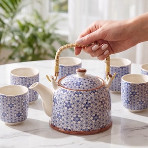 Op de afbeelding: Een keramische theepot met een blauw en wit geometrisch sterpatroon, een bamboe handvat en een bijpassend deksel, vergezeld door vijf kleine bijpassende kopjes. De tekst luidt: Effortless loose-leaf brewing... in one beautifully coordinated set. Built-in strainer. Six matching cups. Always elegant. SPACEENLIGHTENMENT