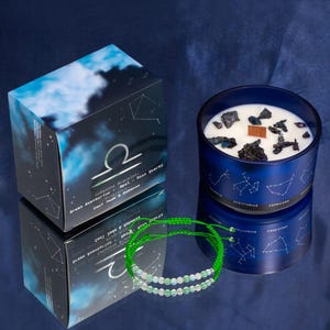Pode incluir: Uma vela azul com pavio de madeira e cristais, uma pulseira de contas verdes e brancas e uma caixa com um símbolo de Libra e texto. A caixa diz "Gemstone Bracelet" e lista pedras. A vela tem constelações do zodíaco.