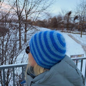 Op de afbeelding: Een blauw en lichtblauw gestreepte gebreide muts. De muts is gemaakt van een zacht, pluizig materiaal en heeft een geribbelde rand. De muts wordt gedragen door een persoon die een zonnebril en een grijze jas draagt, met een besneeuwde buitenachtergrond.