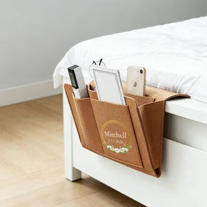 May include: A brown felt bedside organiser with several pockets, suspended from a white bed frame. It contains a remote, a notepad, and a smartphone. The front displays the name "Mitchell" and a floral design.