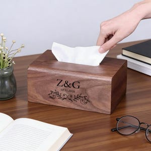 Könnte beinhalten: Eine rechteckige Taschentuchbox aus Holz mit dunkler brauner Oberfläche. Die Box ist mit den Initialen "Z&G", dem Datum "02.11.2026" und einem floralen Design graviert. Weiße Taschentücher sind oben sichtbar.