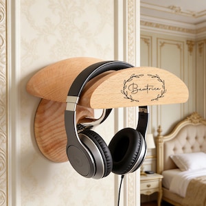 Puede incluir: Soporte de pared para auriculares de madera clara. El soporte tiene un diseño curvo y presenta el nombre "Beatrice" en escritura cursiva dentro de una corona de laurel. Unos auriculares negros y plateados cuelgan del soporte.