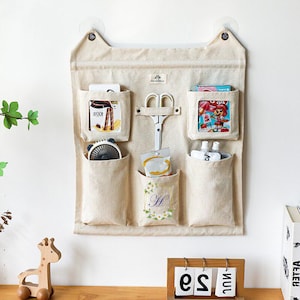 Könnte beinhalten: Beiger Stoff-Wandorganizer mit sechs Taschen und einem Katzenohren-Design. Enthält eine Schere und verschiedene Gegenstände in den Taschen. Der Organizer wird mit Saugnäpfen an der Wand befestigt. Eine Holzgiraffe und ein Kalender befinden sich im Vordergrund.