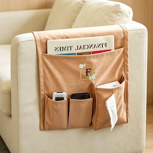 Puede incluir: Un organizador de reposabrazos de sofá de tela color canela con múltiples bolsillos. El organizador tiene un bolsillo grande con un periódico, un bolsillo con un teléfono, un bolsillo con un mando a distancia y un bolsillo con pañuelos. El organizador tiene la letra "F" con detalles florales.