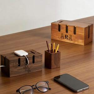 Peut inclure: Des organisateurs de câbles en bois et un porte-stylos sont posés sur un bureau en bois. Les organisateurs ont des fentes pour les câbles et un motif floral décoratif avec la lettre "R". Un bloc de chargement blanc est connecté à l'un des organisateurs. Des lunettes et un smartphone sont également sur le bureau.