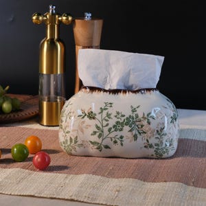 Può includere: Un portatovaglioli in ceramica bianca con un motivo floreale verde e rosa. Il portatovaglioli ha un bordo marrone ed è pieno di fazzoletti bianchi. Un macinapepe dorato e un macinasale in legno sono sullo sfondo.