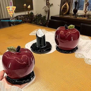 Könnte beinhalten: Drei dekorative Keramikteile auf einem Tisch. Zwei sind glänzende, rot geformte Apfelbehälter mit schwarzen Basen und grünen Blattakzenten. Das dritte ist ein schwarzer Kerzenhalter mit einem weißen Einsatz. Der Tisch ist mit einer gelben Tischdecke bedeckt.