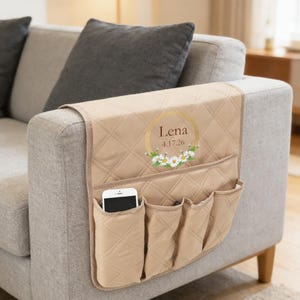 Peut inclure: Organisateur de bras de canapé matelassé beige avec plusieurs poches. Comprend un motif circulaire doré avec le nom "Lena" et la date "4.17.26" ainsi que des accents de marguerites. Un smartphone est visible dans une des poches.