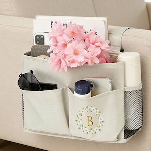 Może przedstawiać: Beżowy organizer na podłokietnik sofy z wieloma kieszeniami. Zawiera książkę, okulary przeciwsłoneczne, telefon, butelkę i bukiet różowych kwiatów. Przednia kieszeń ma wieniec z stokrotek z literą "B".
