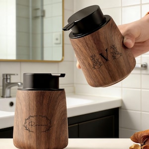 May include: Wooden soap dispenser with a black pump top. The dispenser has a wood grain pattern and a black letter "V" with floral accents. Another dispenser with the name "Rowan" is also visible.