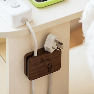 May include: A dark brown wooden cable organizer with a floral design and the letter "g" engraved on the front. A white plug is inserted into the organizer, and a white cable is visible.