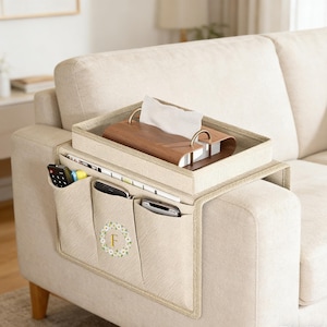 Puede incluir: Organizador de reposabrazos de sofá beige con múltiples bolsillos y una bandeja. El organizador tiene una tela beige claro con una corona floral decorativa alrededor de la letra "F". La bandeja contiene una caja de pañuelos de madera y tiene asas doradas. Los bolsillos contienen mandos a distancia y otros objetos.