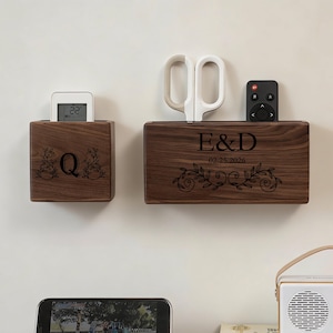 Puede incluir: Dos organizadores de pared de madera con detalles grabados. Uno contiene un pequeño dispositivo digital, el otro un par de tijeras y un mando a distancia. Los organizadores están personalizados con iniciales y una fecha, y presentan diseños florales.