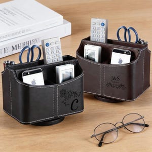 Könnte beinhalten: Zwei Leder-Schreibtisch-Organizer, einer schwarz und einer braun, jeweils mit mehreren Fächern. Der schwarze Organizer hat ein Blumendesign und den Buchstaben "C". Der braune Organizer hat die Initialen "J&S". Beide halten Telefone, Fernbedienungen und Scheren.