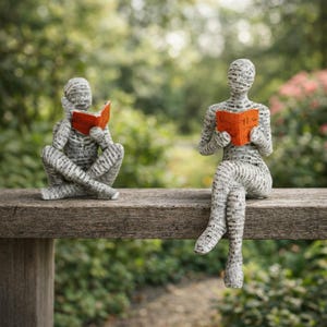 Puede incluir: Dos esculturas de figuras leyendo libros. Las figuras están cubiertas de texto y están sentadas en un banco de madera. Cada figura sostiene un libro naranja. El fondo está borroso con follaje verde.