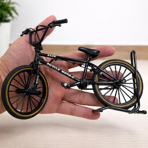 Könnte beinhalten: Ein Miniatur-BMX-Fahrrad in Schwarz mit Reifen mit beigefarbenen Seitenwänden. Auf dem Rahmen des Fahrrads stehen die Wörter "RACE" und "BICYCLE MOTO-X". Das Fahrrad wird von einer Hand gehalten und steht auf einem schwarzen Ständer.