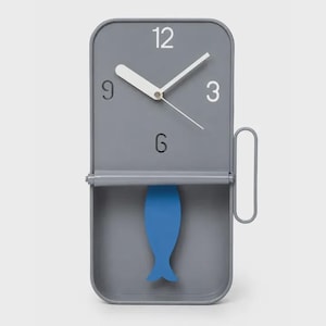 Puede incluir: Un reloj de pared rectangular gris con esquinas redondeadas, que presenta los números blancos 12, 3, 6 y 9. El reloj tiene manecillas blancas para horas y minutos, un segundero blanco fino y un péndulo azul con forma de pez en la parte inferior.