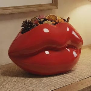 May include: A glossy red ceramic vase shaped like a pair of full lips. The open top of the vase is filled with a decorative arrangement of dried natural elements, including a dark brown pinecone and slices of dried orange.