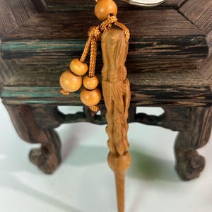 May include: A wooden keychain with a carved figure and a decorative cord with wooden beads. The keychain is attached to a silver metal ring. The figure is light brown and has a detailed design.