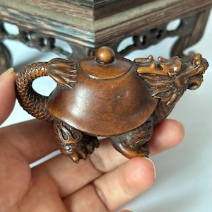 May include: A small, hand-carved brown wooden teapot shaped like a mythical dragon creature. The body of the teapot features a smooth, rounded shell-like centre, while the handle is formed by a textured, scaled tail and the spout is a dragon head.