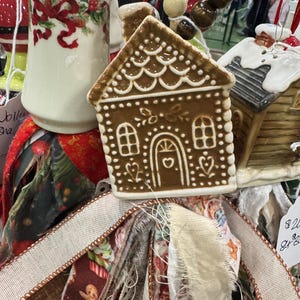 May include: A festive Christmas ornament arrangement featuring a gingerbread house, a ceramic cylinder with red ribbon and holly, and a small house with a snowy roof. The arrangement is set against a backdrop of colorful fabric strips and ribbon.