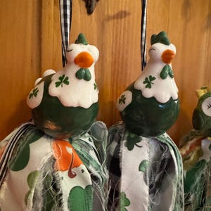 May include: Two ceramic hen ornaments with green and white details, including shamrock accents. The hens have orange beaks and green crests. They are attached to fabric tassels with green shamrock patterns, perfect for St. Patrick's Day.