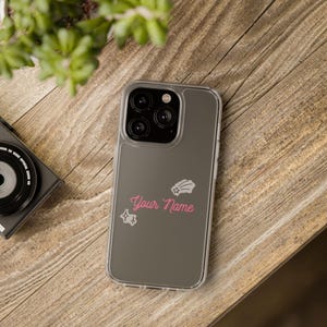 Puede incluir: Funda transparente para teléfono con parte trasera gris que presenta el texto "Your Name" en escritura rosa, junto con gráficos de estrellas y estrellas fugaces. La funda está sobre una superficie de madera.