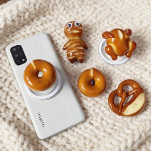 May include: A white smartphone with a donut-shaped phone grip is surrounded by miniature food-themed figurines. The figurines include a pretzel, a roasted chicken, a mummy, and a second donut. The items are arranged on a cream-colored, knitted surface.