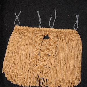 May include: A tan-colored, handmade textile with a fringed bottom and braided center design. The top edge has loops for hanging. The textile is made of yarn and is displayed against a black background. The overall design is symmetrical.