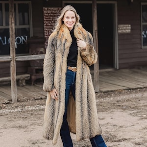 Pu&ograve; includere: Un lungo cappotto di pelliccia marrone e beige con un top nero e jeans blu. Il cappotto ha un ampio colletto e una cintura. Lo sfondo presenta un edificio in legno con la scritta "1000 Saloon".