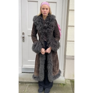 May include: A long, dark brown suede coat featuring thick, shaggy dark gray fur trim along the collar, front opening, cuffs, and hemline. The coat has a structured fit and reaches below the knees, showcasing a textured finish and long sleeves.
