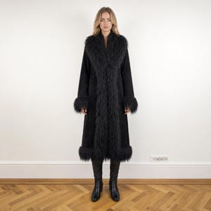 May include: A long black coat featuring a textured, shaggy faux fur trim along the wide collar, the front opening, the sleeve cuffs, and the bottom hem. The coat has a smooth, matte finish and is paired with black knee-high boots.