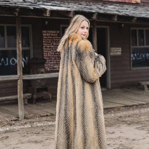 Pu&ograve; includere: Un cappotto lungo in finta pelliccia con un motivo a spina di pesce nei toni del marrone, beige e nero. Il cappotto &egrave; indossato con jeans blu scuro e stivali da cowboy marroni. Lo sfondo presenta un edificio in legno con la scritta "1000 Saloon".