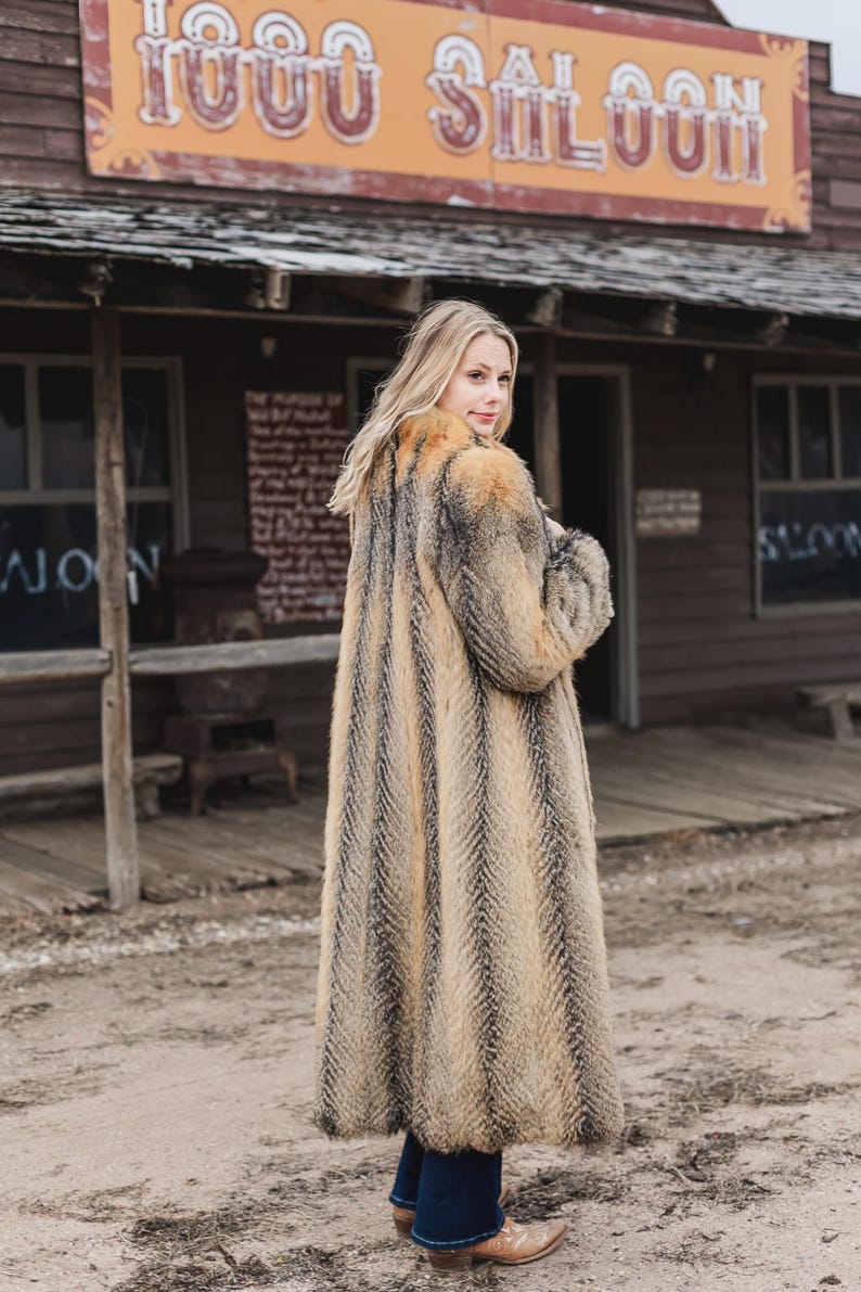 Pu&ograve; includere: Un cappotto lungo in finta pelliccia con un motivo a spina di pesce nei toni del marrone, beige e nero. Il cappotto &egrave; indossato con jeans blu scuro e stivali da cowboy marroni. Lo sfondo presenta un edificio in legno con la scritta "1000 Saloon".