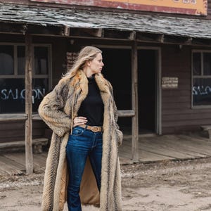Pu&ograve; includere: Un lungo cappotto di pelliccia marrone e beige, indossato sopra un top nero e jeans blu. Il cappotto ha un motivo a righe verticali. Lo sfondo presenta un edificio in legno con l'insegna "1000 Saloon".