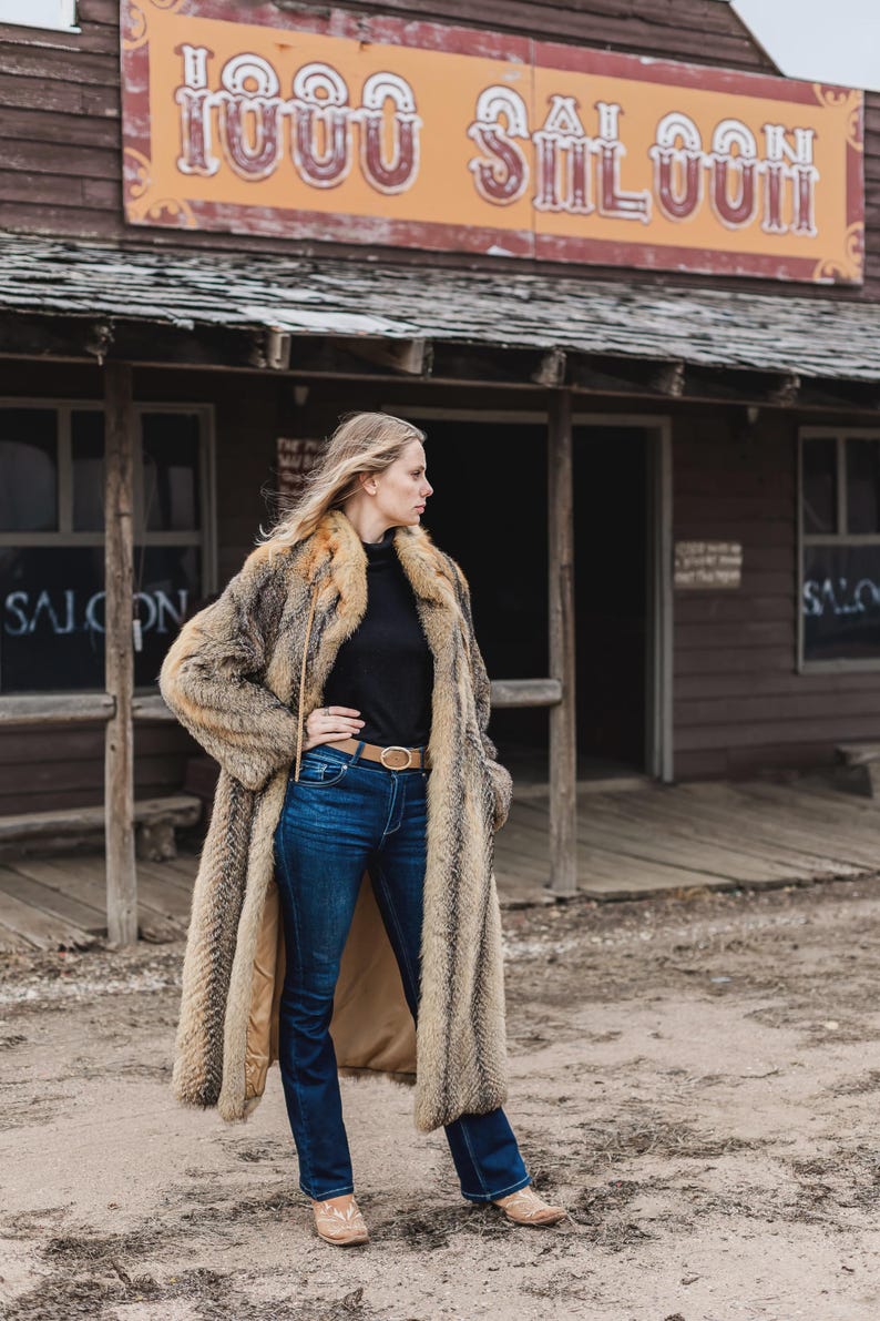 Pu&ograve; includere: Un lungo cappotto di pelliccia marrone e beige, indossato sopra un top nero e jeans blu. Il cappotto ha un motivo a righe verticali. Lo sfondo presenta un edificio in legno con l'insegna "1000 Saloon".