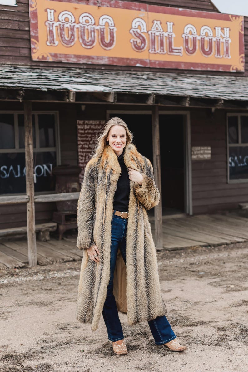 Pu&ograve; includere: Un lungo cappotto di pelliccia marrone e beige con un top nero e jeans blu. Il cappotto ha un ampio colletto e una cintura. Lo sfondo presenta un edificio in legno con la scritta "1000 Saloon".