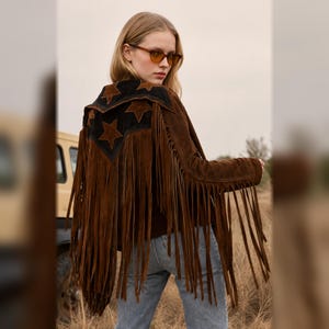 Könnte beinhalten: Eine braune Wildlederjacke mit langen Fransen an den Ärmeln und am Rücken. Das Rückenteil hat eine schwarze Passe, die mit braunen sternförmigen Applikationen verziert ist. Die Jacke wird mit einer hellblauen Jeans und einer bernsteinfarbenen Brille getragen.
