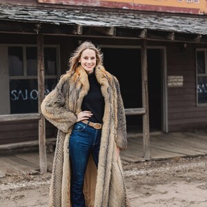 Pu&ograve; includere: Un lungo cappotto di pelliccia nei toni del marrone e beige, indossato sopra un dolcevita nero e jeans blu. Il cappotto ha un aspetto lussuoso. Lo sfondo presenta un edificio in legno con la scritta "1000 Saloon".