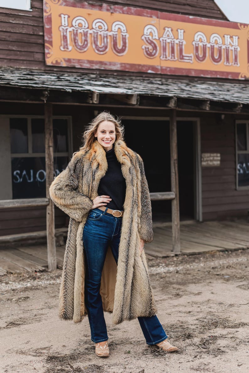Pu&ograve; includere: Un lungo cappotto di pelliccia nei toni del marrone e beige, indossato sopra un dolcevita nero e jeans blu. Il cappotto ha un aspetto lussuoso. Lo sfondo presenta un edificio in legno con la scritta "1000 Saloon".