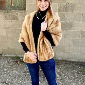 May include: A tan-colored fur wrap with a soft, textured appearance. The wrap is draped over the shoulders, creating a luxurious look. The person is wearing a black turtleneck and blue jeans.