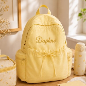 May include: A pale yellow backpack featuring the name Daphne embroidered in cursive script on the front pocket, which includes a decorative drawstring bow. The backpack has side pockets with zippers and a top handle for carrying.