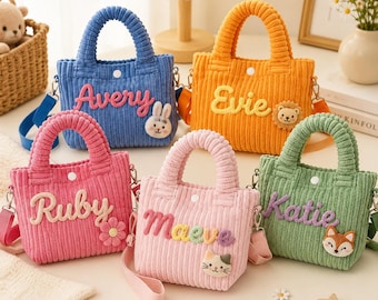 May include: Five corduroy tote bags in blue, orange, pink, light pink, and green. Each bag features a white snap closure, a shoulder strap, and a name embroidered in cursive: Avery with a rabbit, Evie with a lion, Ruby with a flower, Maeve with a cat, and Katie with a fox.