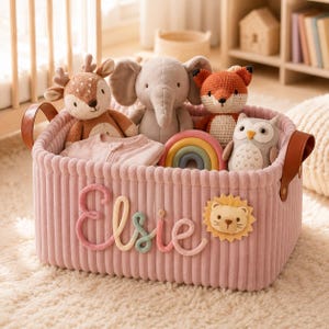 Op de afbeelding: Een roze corduroy opbergmand met bruine lederen handvatten, voorzien van de naam Elsie in pastelkleurige garenletters en een leeuwenembleem. Binnenin zitten knuffels zoals een hert, olifant, vos en uil, plus een regenboogspeeltje en een roze kledingstuk.