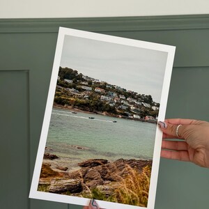 May include: A framed print depicting a coastal scene with a town on a hillside. The image shows a tranquil bay with boats, rocky shores, and golden grasses in the foreground. The print is held against a green wall.