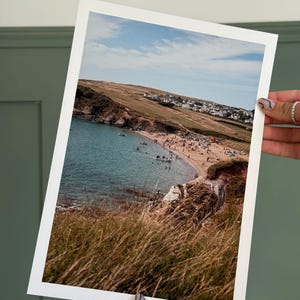 May include: A framed print depicting a beach scene with a sandy shore, clear turquoise water, and a hillside with buildings. The sky is blue with clouds. The foreground features tall, dry grass.