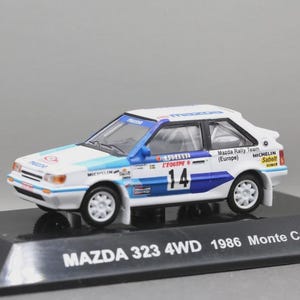 Op de afbeelding: Een wit en blauw Mazda 323 4WD 1986 Monte Carlo modelauto. De miniatuurauto heeft race-decals, het nummer 14 en sponsorlogo's. Het model wordt gepresenteerd op een zwarte basis.